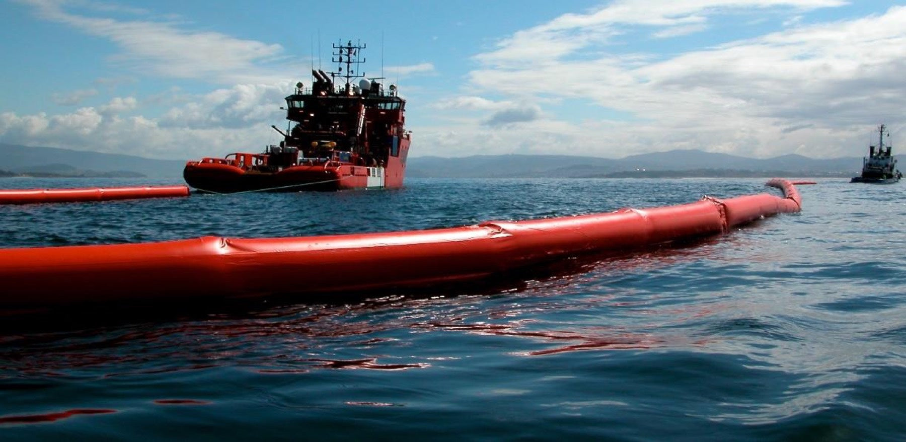 Barrera de contención naranja desplegada en el mar junto a un buque